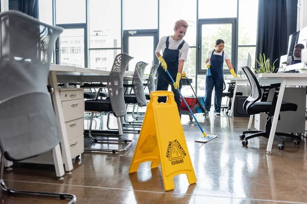 woman in overalls washing floor with mop near attention board and bi-racial colleague