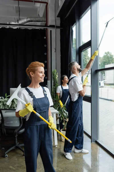 Lächelnde Frau Uniform Hält Scheibenwischer Während Interrassische Kollegen Bürofenster Waschen — Stockfoto