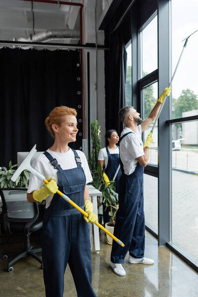 smiling woman in uniform holding window wiper while interracial colleagues washing office windows