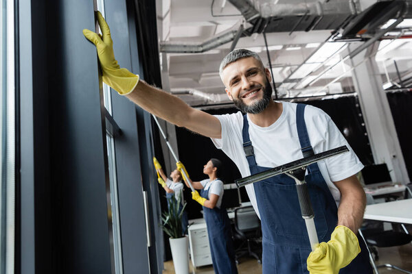 happy bearded man with window wiper smiling at camera near multiethnic women on blurred background