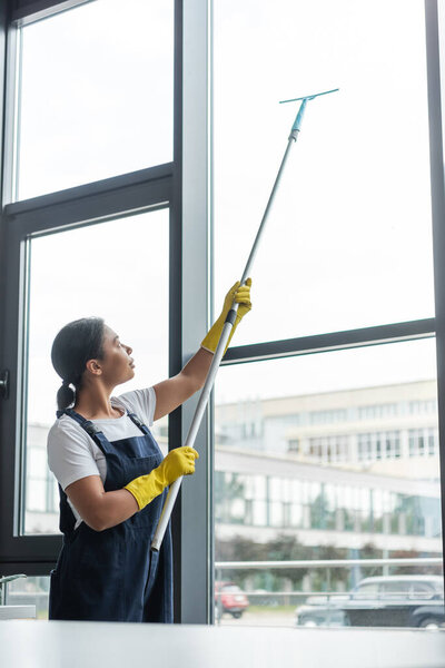 bi-racial woman in overalls and rubber gloves cleaning large windows in office