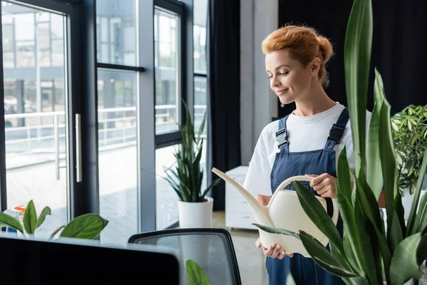 smiling woman in overalls holding watering can near green plant in office