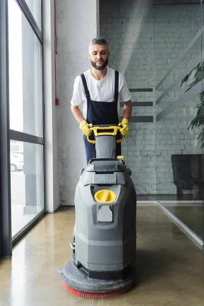 bearded man in overalls cleaning floor with electrical scrubber machine