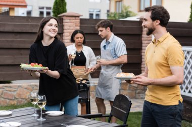 Positive people holding food near wine and interracial friends during picnic in backyard  clipart