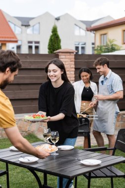 Smiling woman holding fresh salad near multiethnic friends during picnic in backyard  clipart