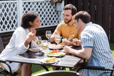 Gülümseyen adam, arka bahçede yemek ve ırklar arası dostların yanındaki akıllı telefonu işaret ediyor. 