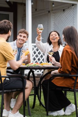 happy bi-racial woman raising glass of wine near friends during bbq party in backyard  clipart