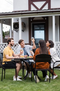 happy man raising glass of wine near interracial friends during bbq party on backyard  clipart