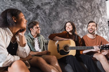 Modern oturma odasındaki koltukta oturan çok ırklı arkadaşlarının yanında akustik gitar tutan neşeli bir kadın. 