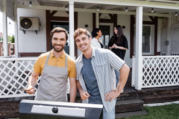 Sonrientes Amigos Mirando Cámara Cerca Parrilla Borrosa Interracial Mujeres Patio — Foto de Stock