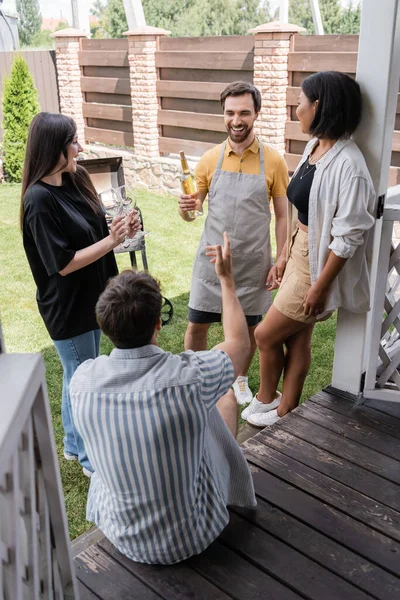 Hombre Sonriente Delantal Sosteniendo Botella Vino Cerca Amigos Multiétnicos Parrilla — Foto de Stock