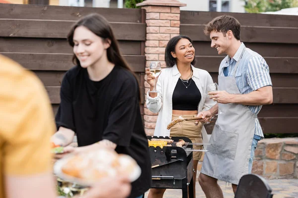 Smiling bi-racial woman holding wine while friend cooking sausages on grill outdoors
