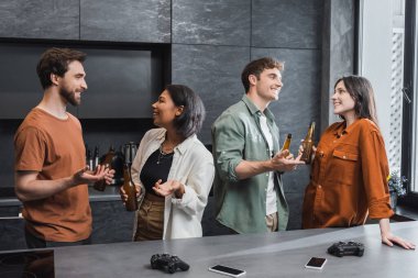 KYIV, UKRAINE - 26 Temmuz 2022: Mutfak tezgahının yanında bira şişeleri ve joysticklerle sohbet eden mutlu ırklar arası arkadaşlar 