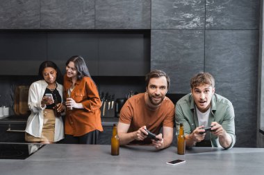 KYIV, UKRAINE - 26 Temmuz 2022: Mutfakta akıllı telefon kullanan ırklar arası kadınların yakınında video oyunu oynayan mutlu erkekler 