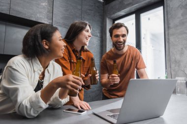 Mutlu çoklu etnik arkadaşlar dizüstü bilgisayarın yanında bira şişeleri ve mutfak atölyesinde boş ekranlı akıllı telefon tutuyorlar.