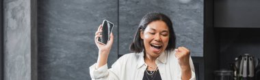 Heyecanlı çift ırklı kadın elinde akıllı telefonuyla mutfakta boş ekran ve afiş tutuyor. 