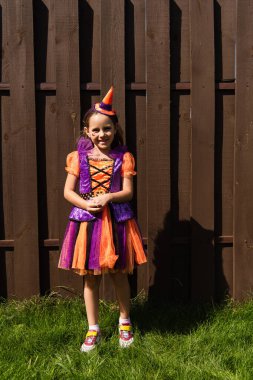 full length of girl in clown costume and party cap smiling near wooden fence clipart