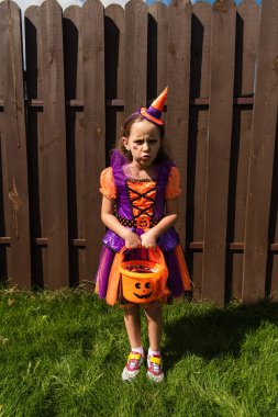 offended girl in clown costume holding halloween bucket with sweets and looking at camera clipart