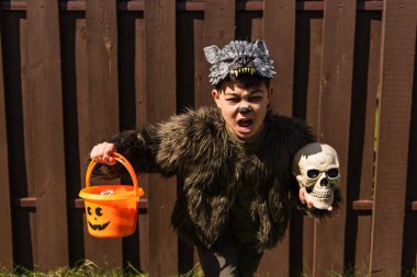 asian boy in werewolf costume running with skull and trick or treat bucket while scary screaming outdoors clipart