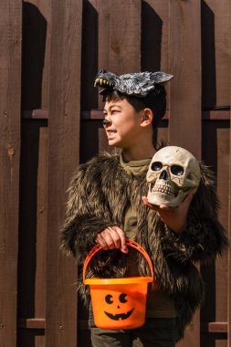 smiling asian boy in werewolf costume looking away while standing with trick or treat bucket and skull clipart