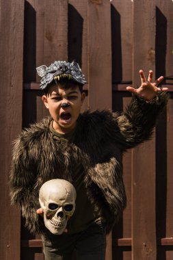 asian boy in werewolf costume holding skull while showing angry grimace and frightening gesture clipart
