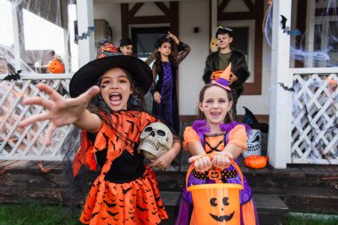 girl in witch costume screaming with outstretched hand near smiling friend with trick or treat bucket clipart