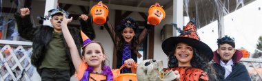 cheerful multiethnic friends in halloween costumes with trick or treat buckets near cottage, banner clipart