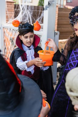 Asian boy in vampire costume holding bucket near friends during halloween celebration outdoors clipart