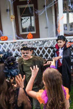 Asian boy in halloween costume grimacing near blurred friends in backyard  clipart