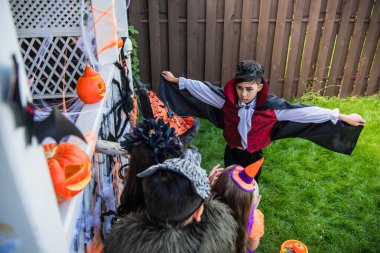 Overhead view of asian boy in halloween vampire costume standing near friends and decor in backyard clipart