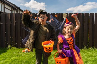 Multiethnic children in halloween costumes holding buckets and grimacing at camera in backyard  clipart
