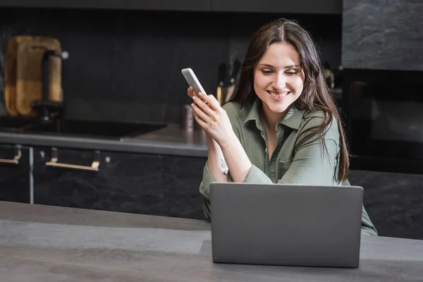 Cheerful Young Woman Green Shirt Holding Smartphone Looking Laptop — Stock Photo, Image