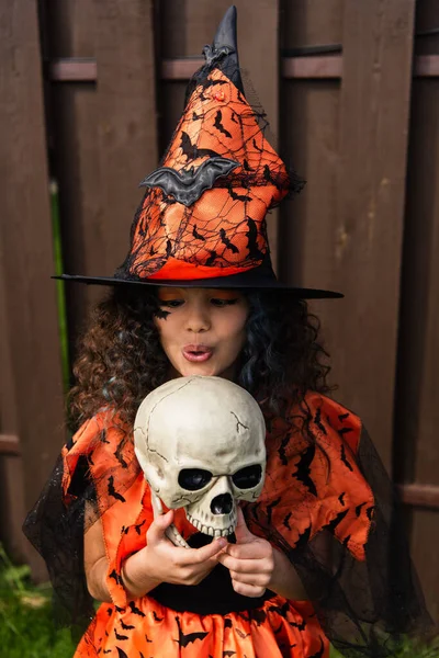curly girl in witch hat pouting lips near spooky skull outdoors