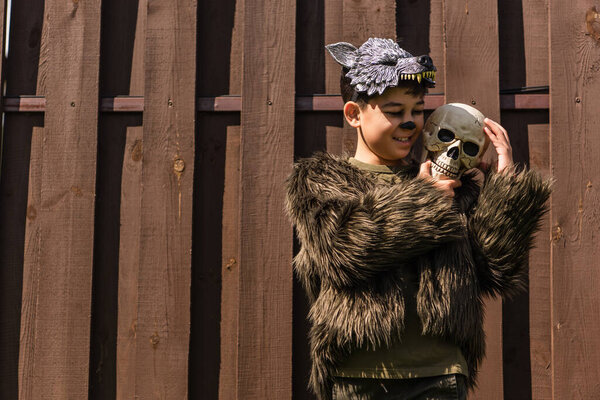 happy asian boy in halloween werewolf costume embracing spooky skull near wooden fence