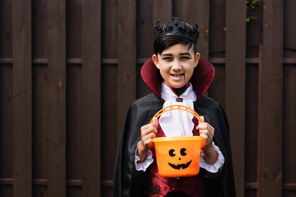 asian boy in vampire king costume holding halloween bucket and looking at camera
