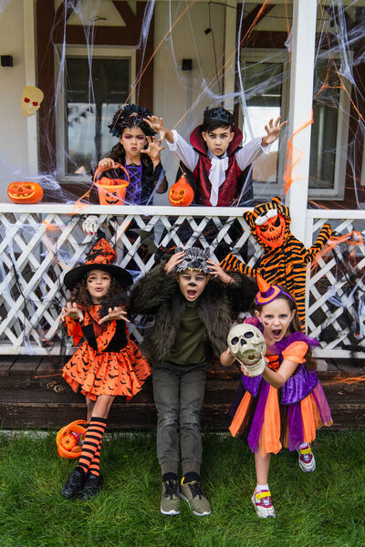 Multiethnic kids in halloween costumes grimacing near jack o lanterns in backyard 