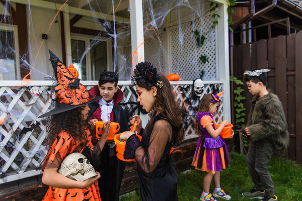 Preteen girls in halloween costumes holding skull and bucket near multiethnic friends in backyard of house 