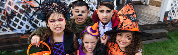 Interracial preteen friends in halloween costumes grimacing at camera outdoors, banner 