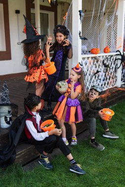 asian boys sitting with halloween buckets near girls in witch costumes scaring each other clipart