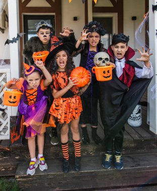 Multiethnic children in halloween costumes holding buckets and grimacing at camera near house clipart