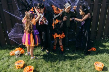 Cadılar Bayramı kostümlü çoklu etnik çocuklar arka bahçedeki dekorun yanında birbirlerini korkutuyorlar.
