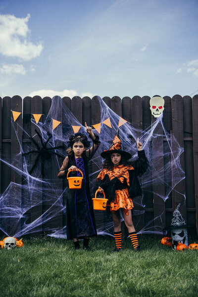 Girls in halloween costumes holding buckets and pointing with fingers near decor on fence outdoors