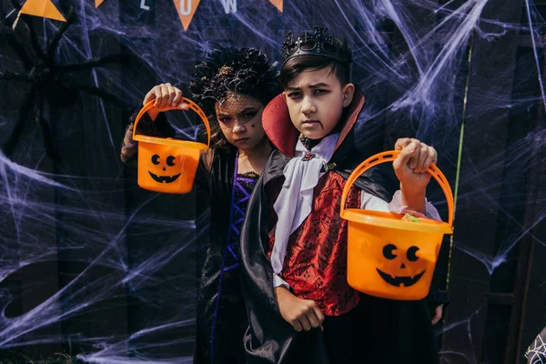 Niños Serios Multiétnicos Trajes Halloween Sosteniendo Cubos Cerca Decoración Patio — Foto de Stock