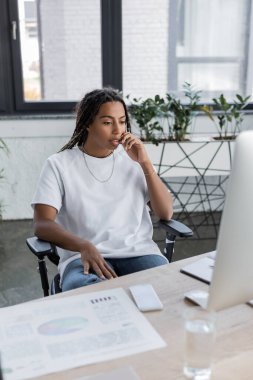 İş yerindeki akıllı telefonun yanındaki bilgisayara günlük kıyafetlerle bakan Afrikalı Amerikalı iş kadını. 