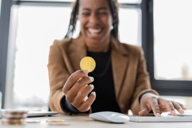 KYIV, UKRAINE - 27 Nisan 2022: Ofiste bilgisayar klavyesinin yanında Bitcoin tutan bulanık Afrikalı Amerikalı iş kadını 