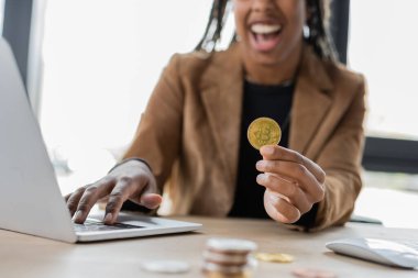 KYIV, UKRAINE - 27 Nisan 2022: Ofiste bilgisayarın yanında bitcoin tutan Afrikalı Amerikalı iş kadınının bulanık görüntüsü 