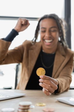 KYIV, UKRAINE - 27 Nisan 2022: Ofiste bilgisayarın yanında Bitcoin tutan heyecanlı Afrikalı Amerikalı iş kadını 