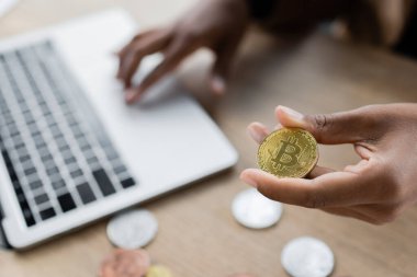KYIV, UKRAINE - 27 Nisan 2022: Afrika kökenli Amerikalı iş kadını altın bitcoin tutarken ve ofiste dizüstü bilgisayar kullanırken görüntülendi 