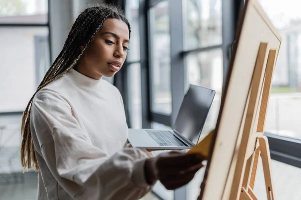 African american businesswoman holding laptop and looking at board in office