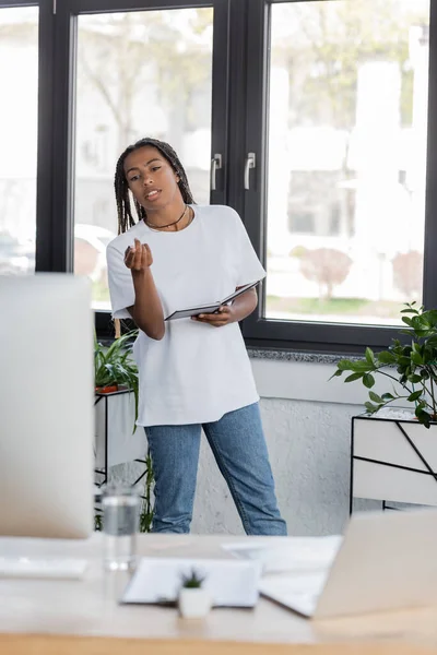 African american businesswoman in casual clothes holding notebook and pointing at computer in office